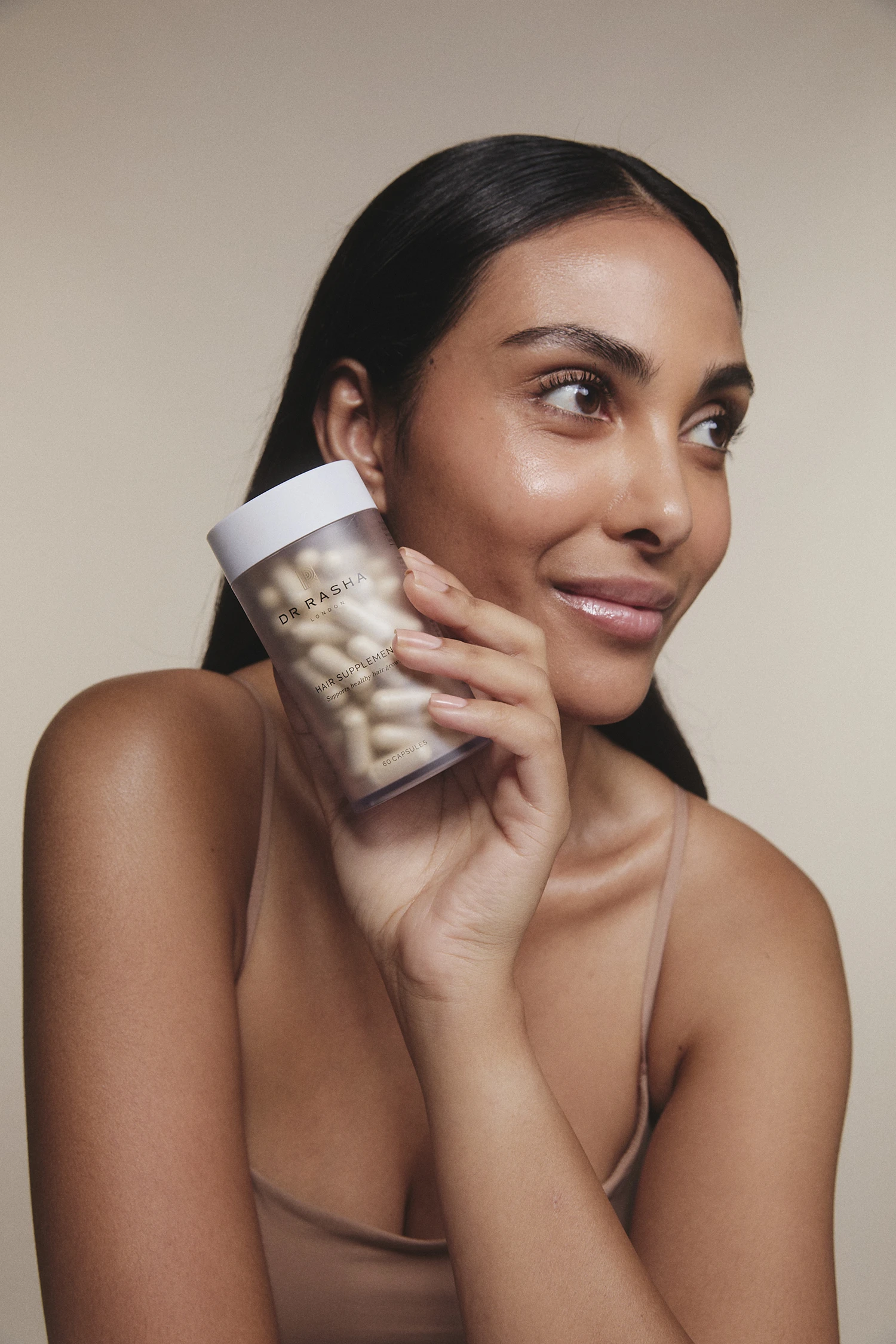 Smiling woman with long dark hair holds a bottle of Dr Rasha hair supplements near her face.