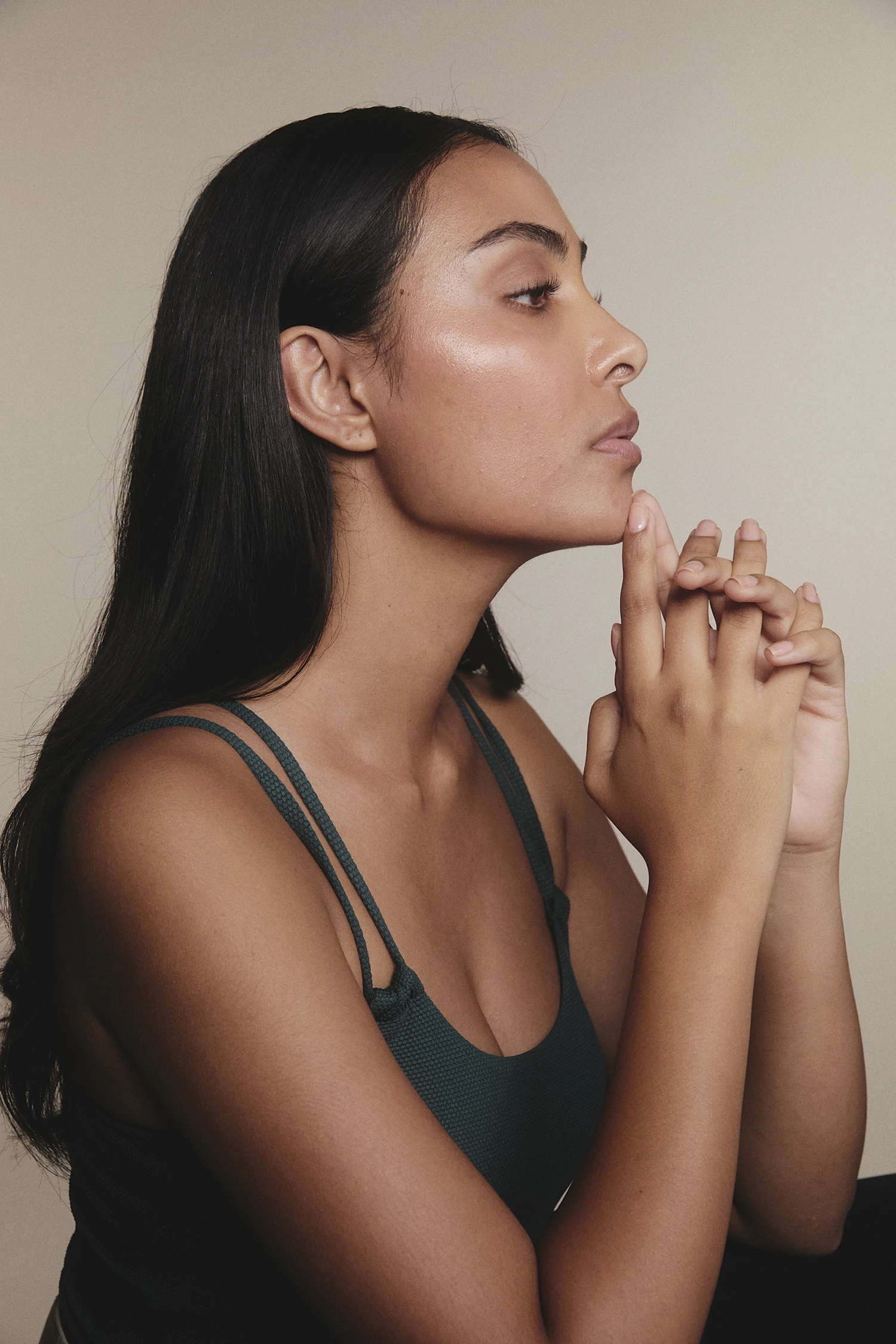 Side profile of a woman with long dark hair wearing a sleeveless top, hands clasped near her chin.