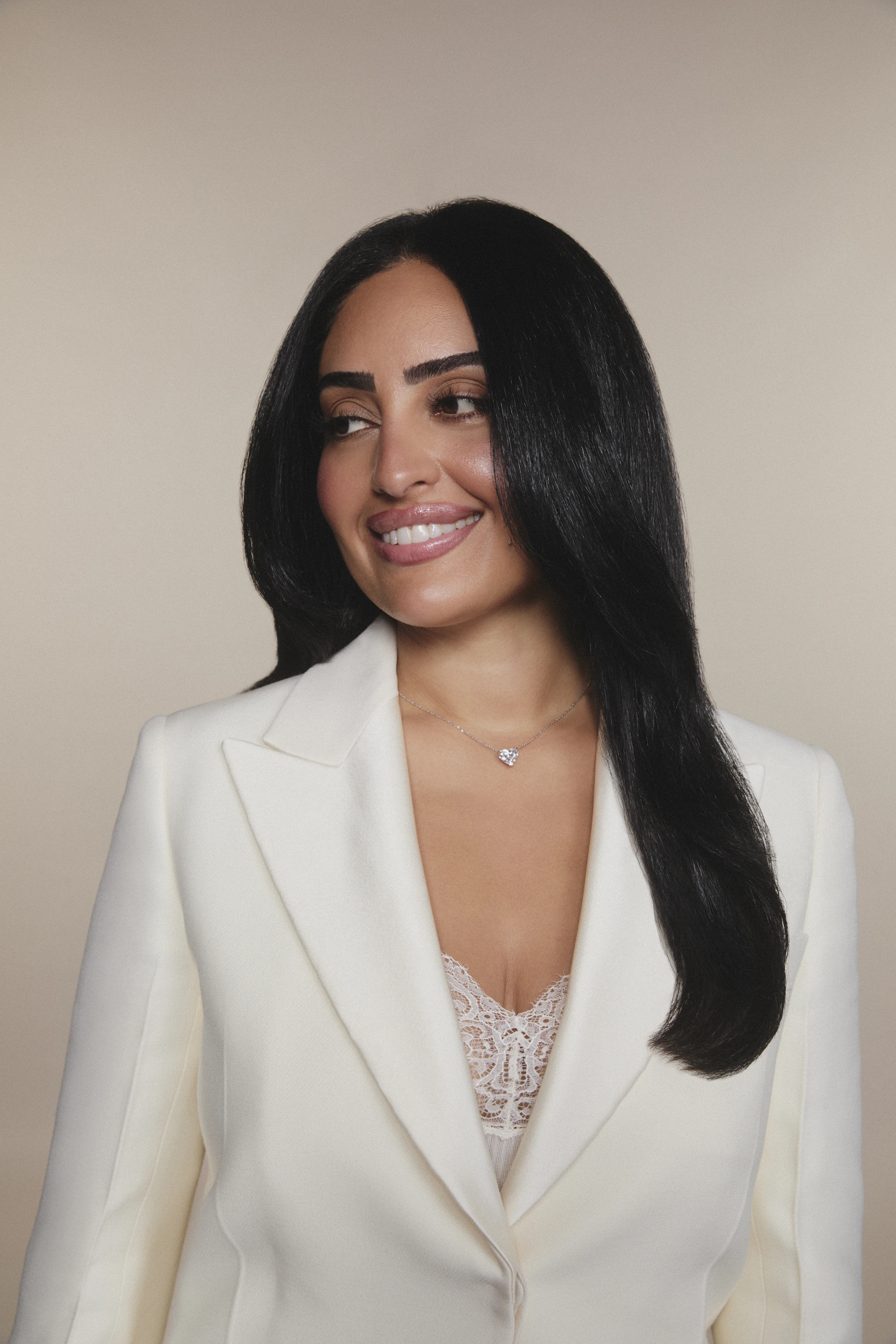 Woman with long black hair wearing a white blazer and lace top, smiling and looking left.
