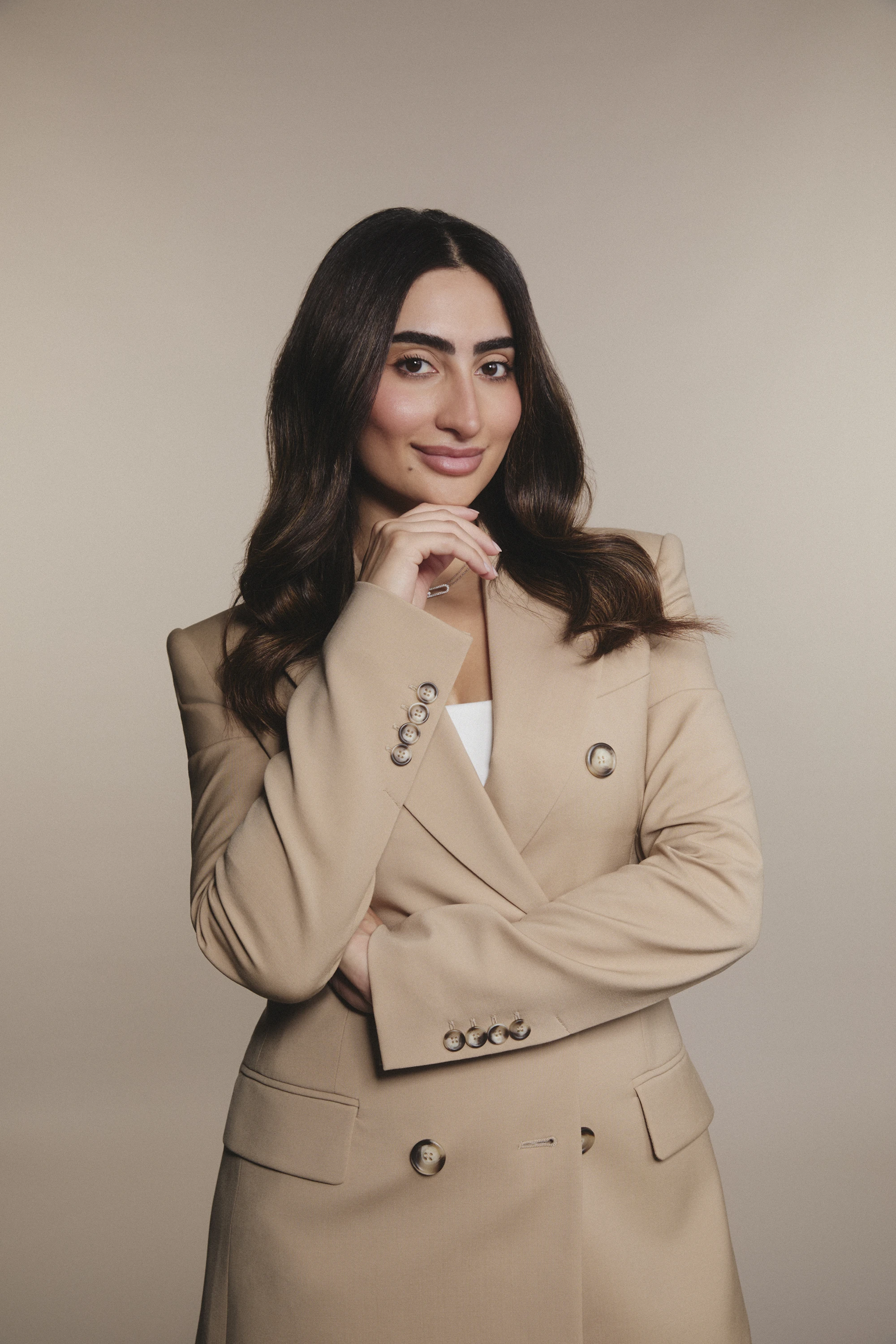 Confident woman with long dark hair and beige blazer, smiling with hand near chin against neutral background.