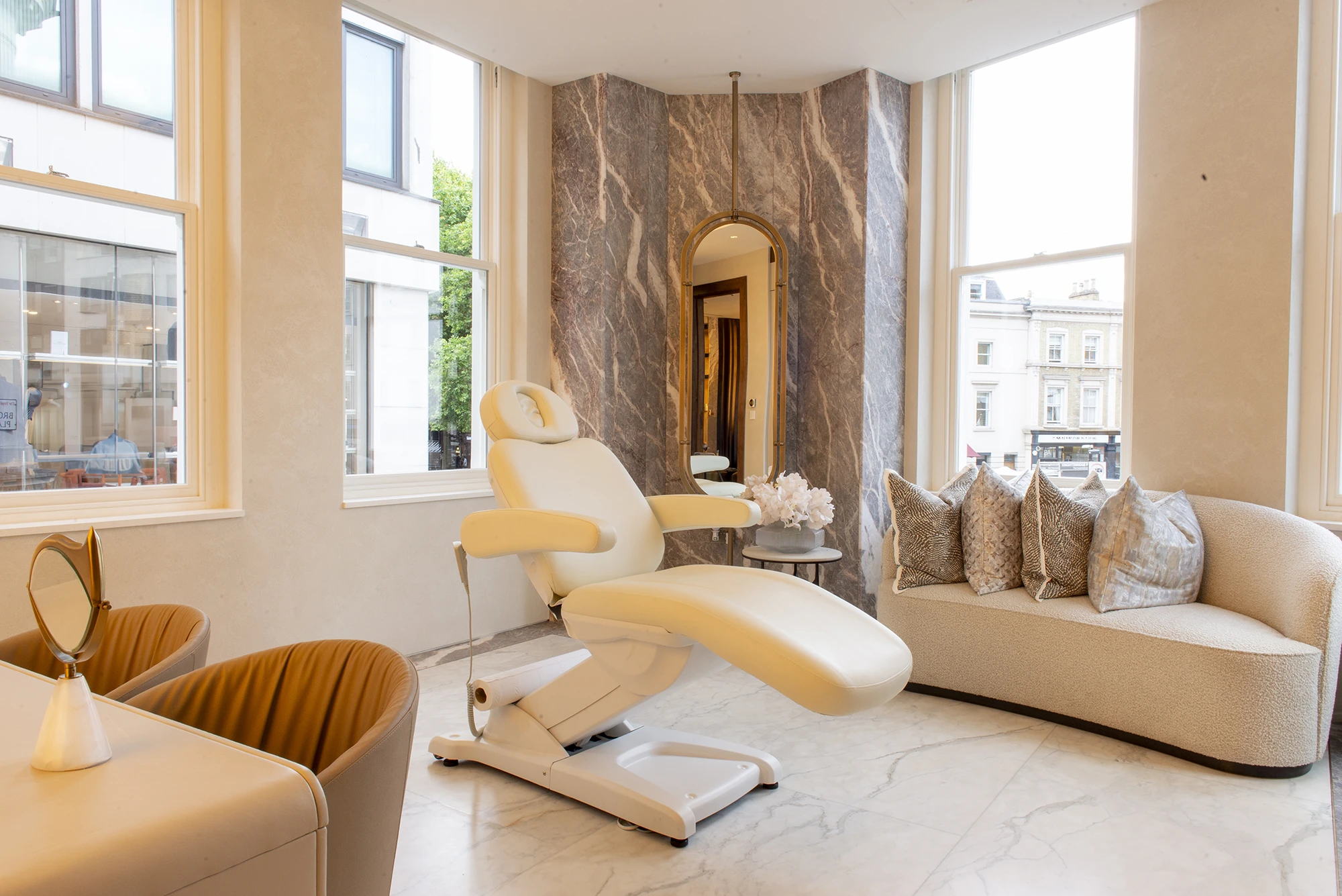 Modern spa room with reclining chair, curved sofa with pillows, arched mirror, and large windows.