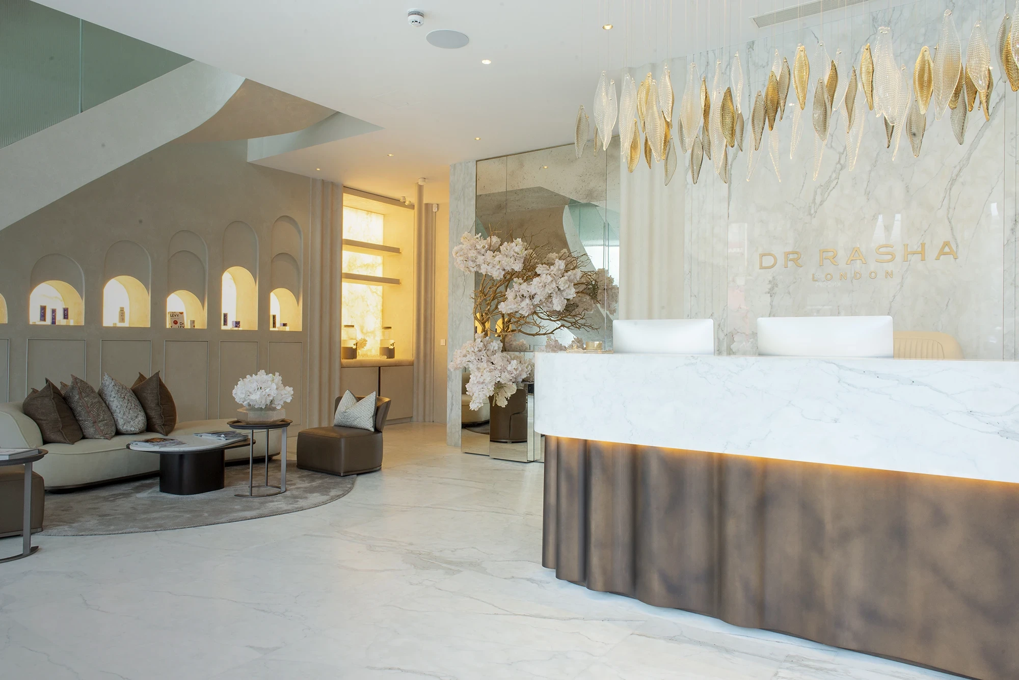Modern clinic reception with marble desk, white chairs, brown sofa with pillows, and decorative leaves overhead.