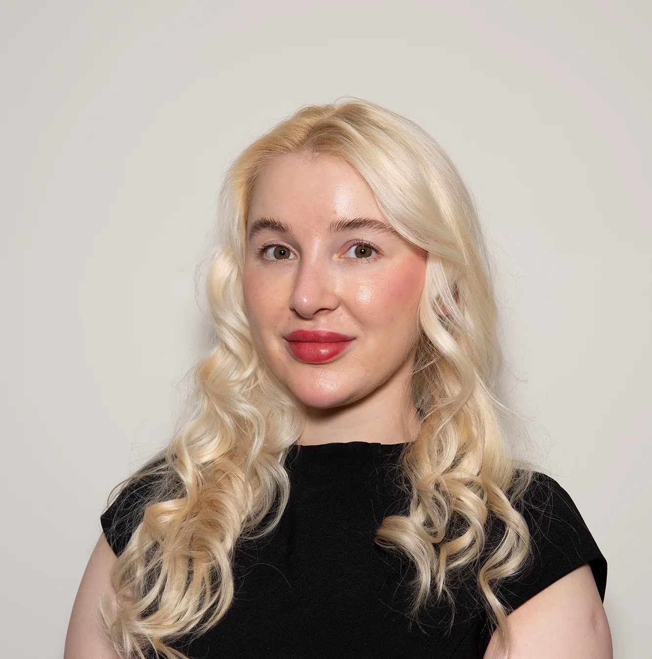 Blonde woman with long curly hair and red lipstick wearing a black top against a plain background.