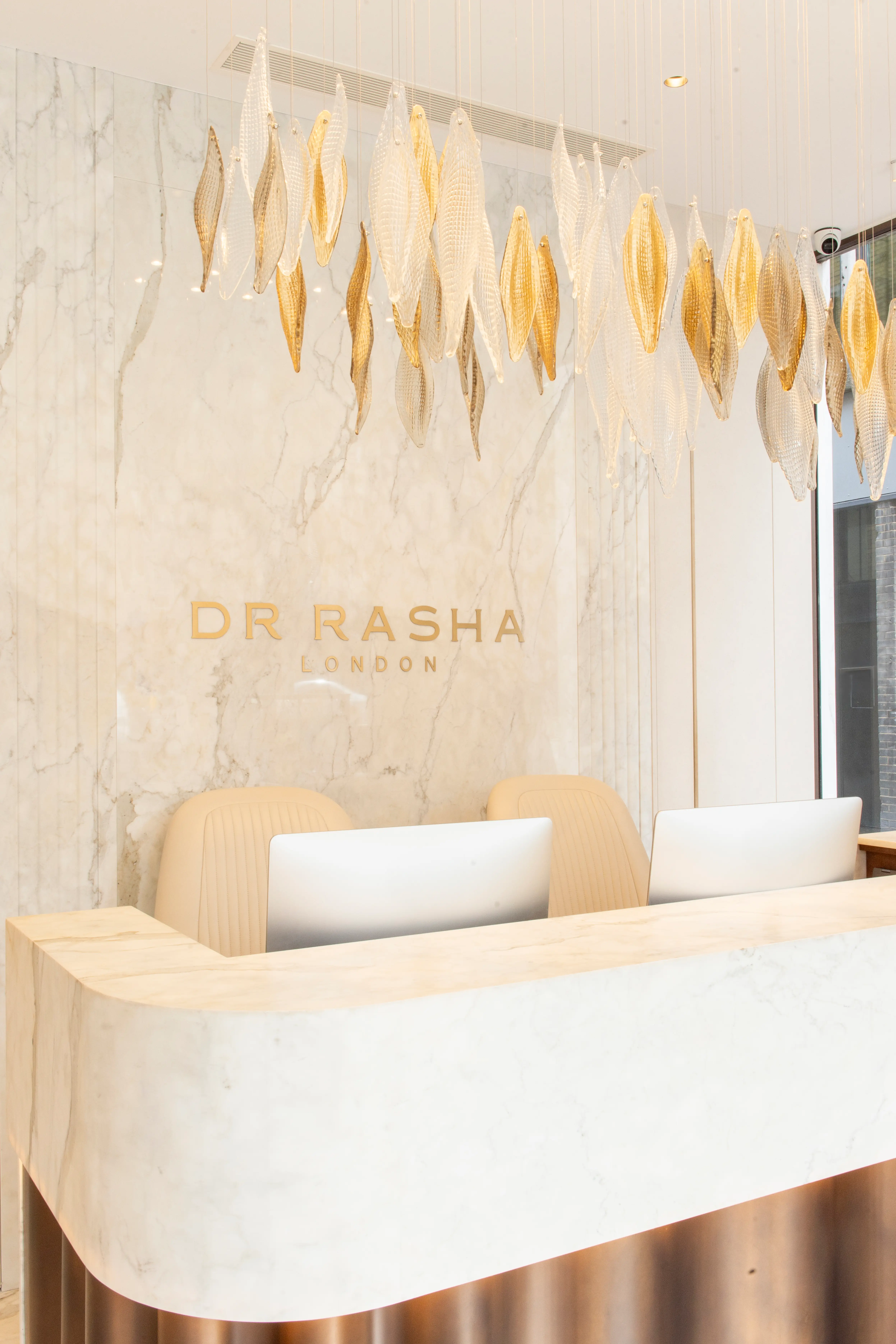 Modern reception area with white marble desk, beige chairs, gold pendant art, and Dr Rasha London sign.