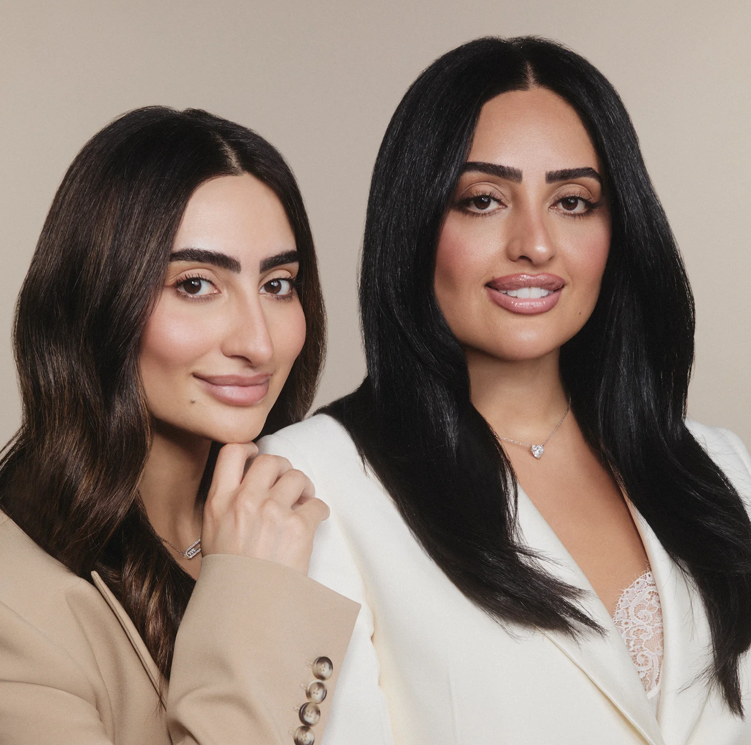 Two women with dark hair wear beige and white blazers, smiling against a plain background.