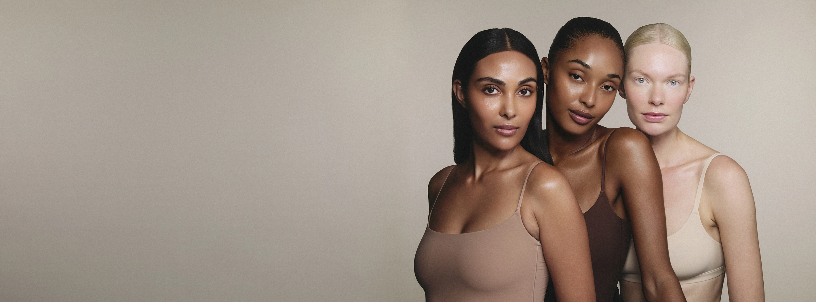 Three women of diverse skin tones wearing neutral-toned tank tops against a beige background.