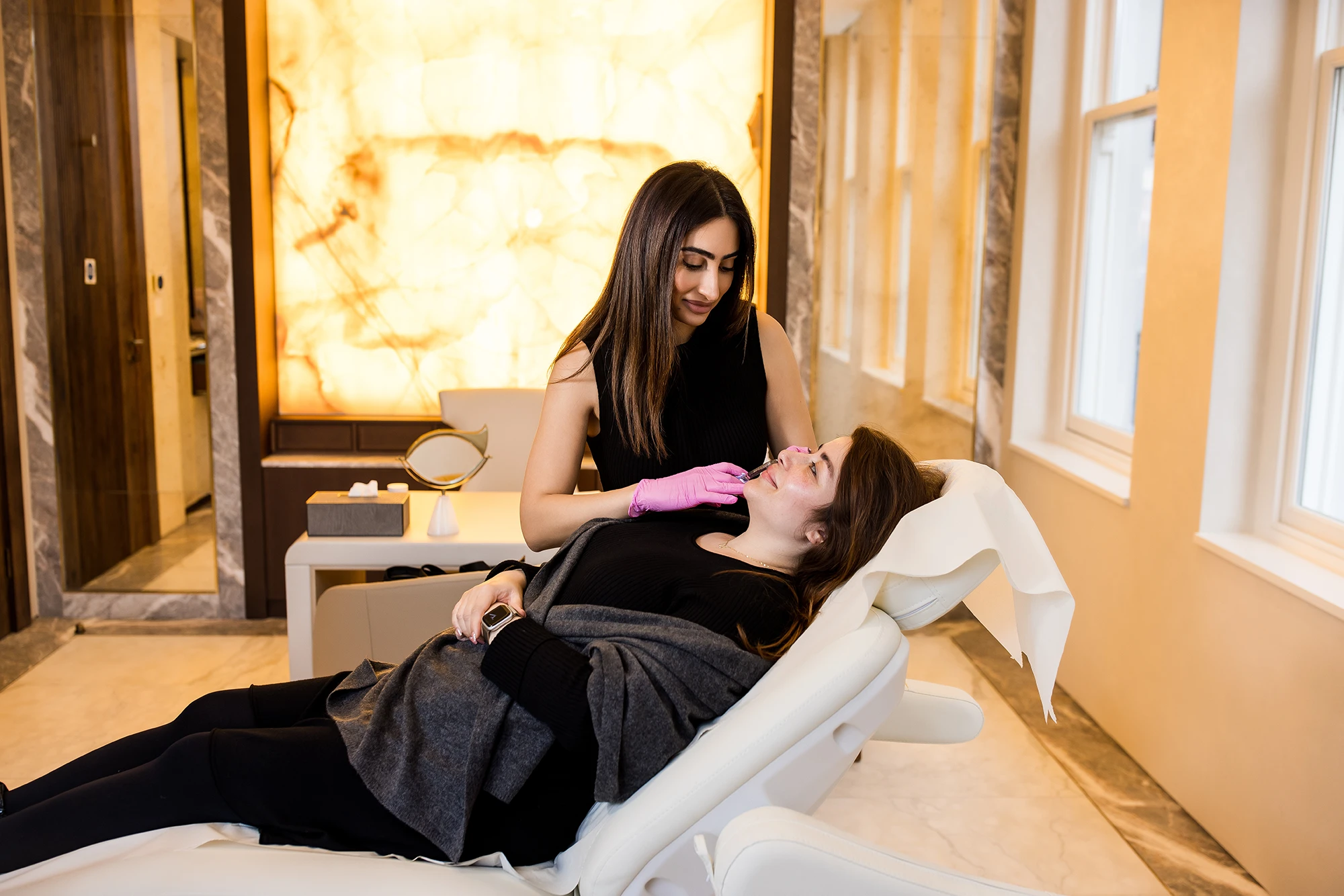 Esthetician wearing pink gloves performs facial treatment on a woman reclining in a spa chair.