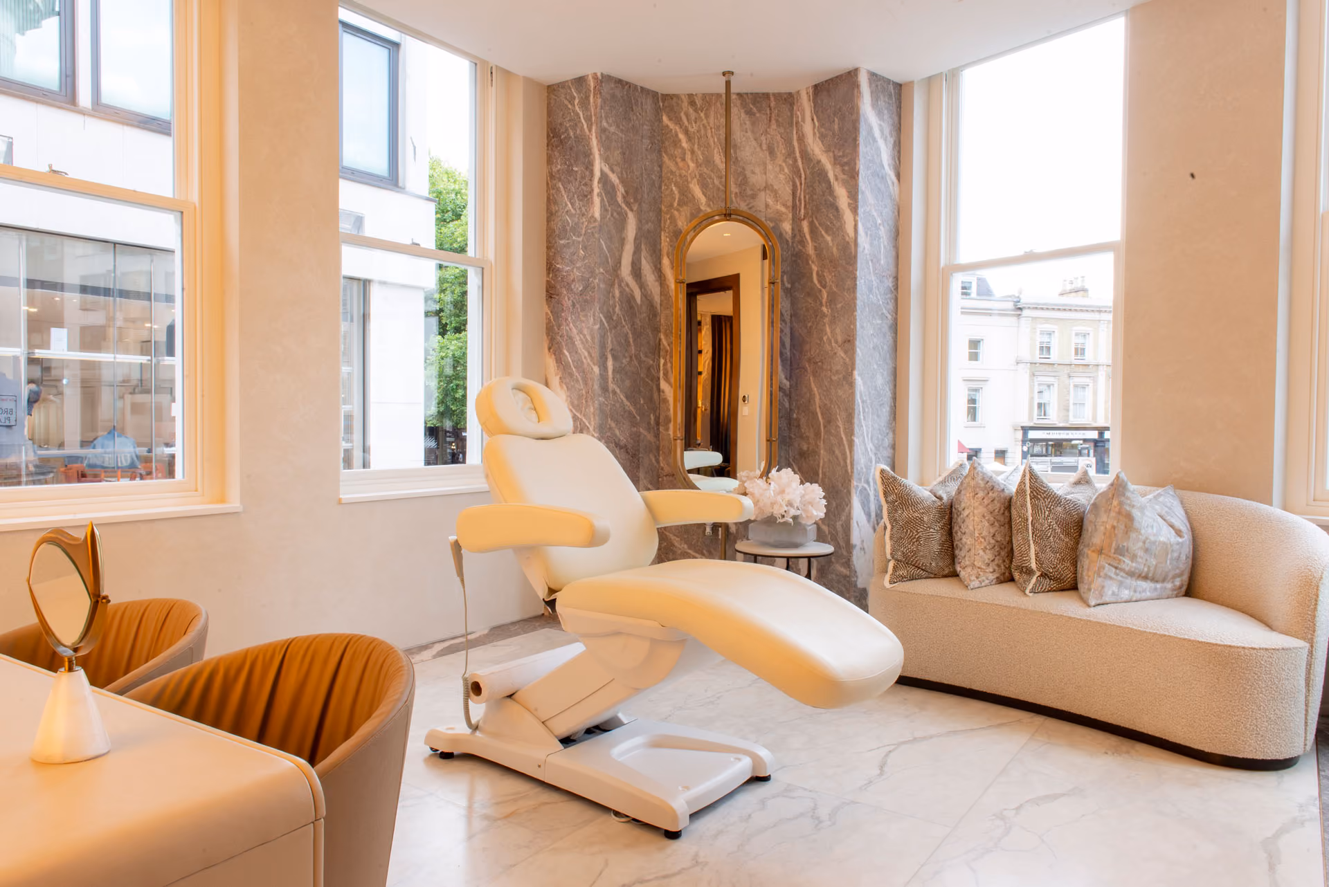 Bright clinic room with white treatment chair, curved sofa with pillows, large windows, and a tall mirror.