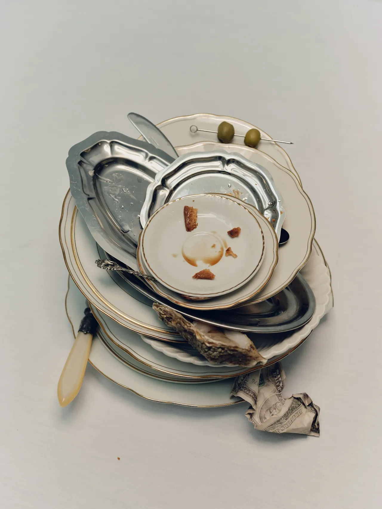  A stack of ornate white and silver plates with food remnants, a butter knife with a cream handle, olives on a cocktail pick, an oyster shell, and a crumpled dollar bill tucked beneath the dishes against a plain white background.