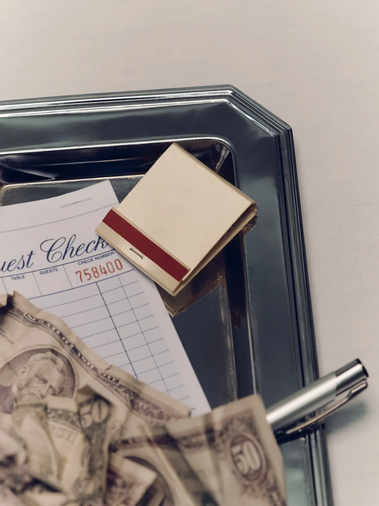 A silver serving tray holds a restaurant guest check, a small beige matchbook with a red stripe, crumpled paper bills, and a metallic pen. The objects are arranged against a pale neutral background.