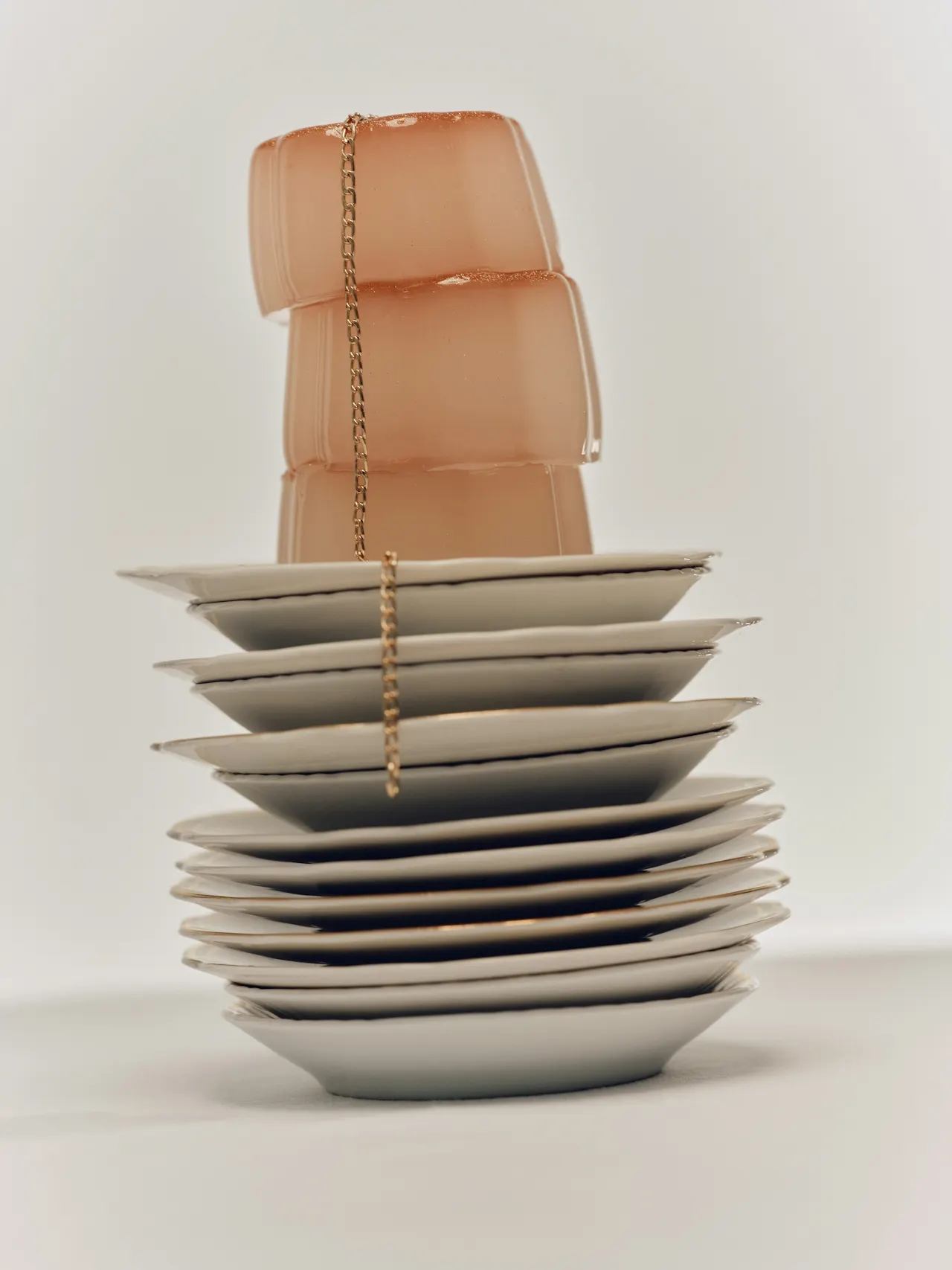 A tall stack of white plates topped with translucent peach gelatin cubes, with a delicate gold chain draped vertically over the stack, set against a minimalist white background.