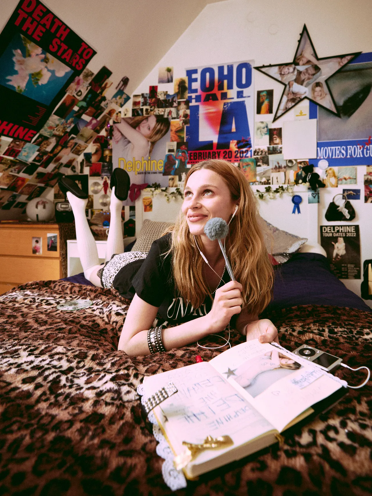 A young woman lies on her stomach across a bed, smiling and looking upward while holding a fuzzy microphone near her face as if recording or speaking into it. An open notebook and music player sit beside her, and the bedroom walls are densely covered with colorful posters, photographs, and magazine clippings.