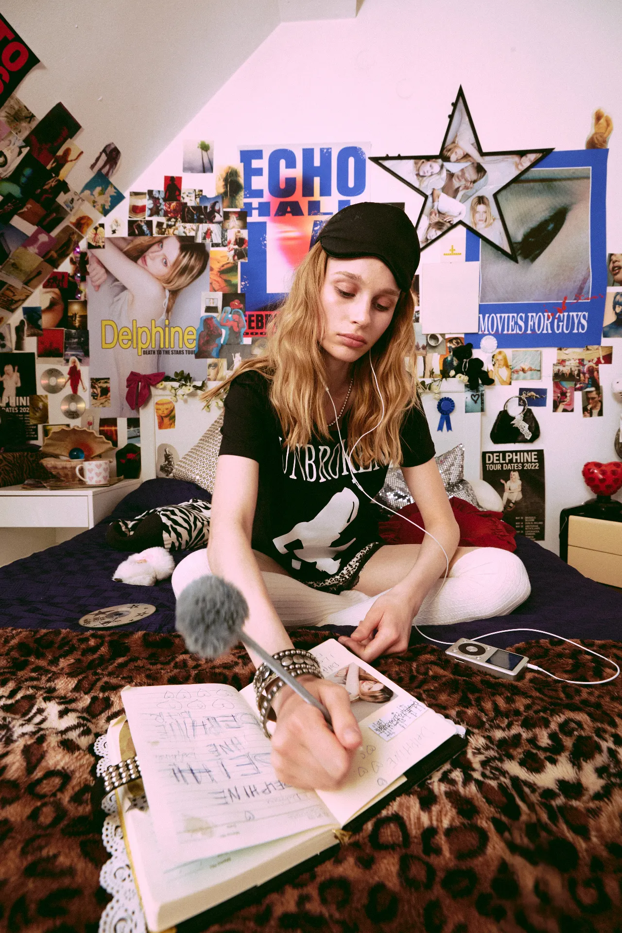 A young woman wearing a black T-shirt, sleep mask, and headphones sits on a bed writing in a notebook. The bedroom is covered in colorful posters, photographs, CDs, and magazine clippings, with an iPod beside her and a fuzzy microphone in the foreground.