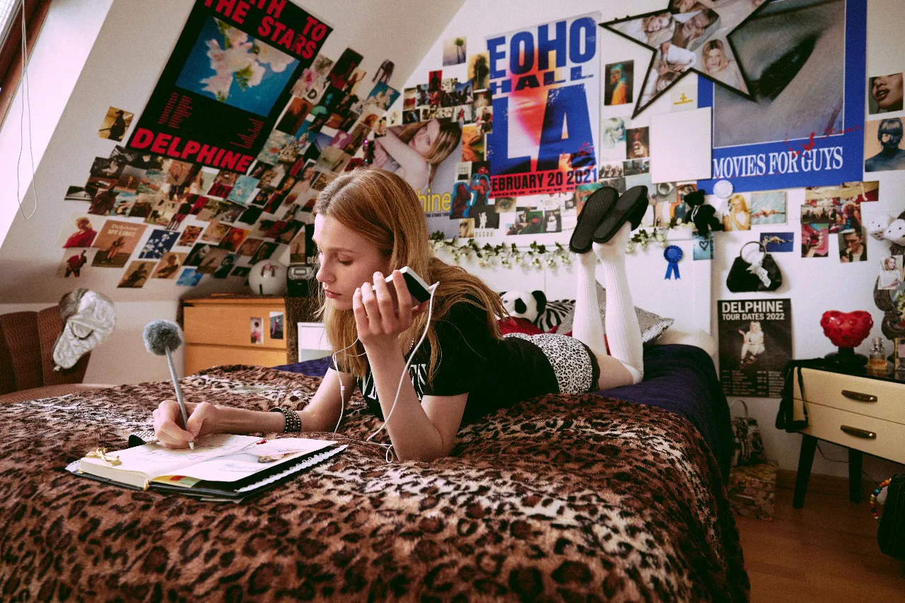 A young woman lies on her stomach across a bed, smiling and looking upward while holding a fuzzy microphone near her face as if recording or speaking into it. An open notebook and music player sit beside her, and the bedroom walls are densely covered with colorful posters, photographs, and magazine clippings.