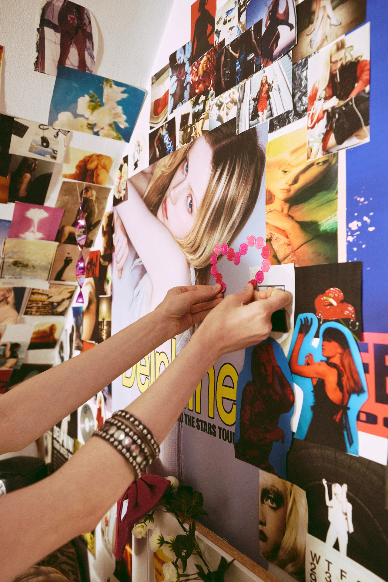 A close-up of a woman’s hands attaching a small pink heart-shaped decoration to a bedroom wall covered in photographs, posters, and magazine clippings. The collage includes portraits, fashion imagery, flowers, and colorful graphics layered closely together.
