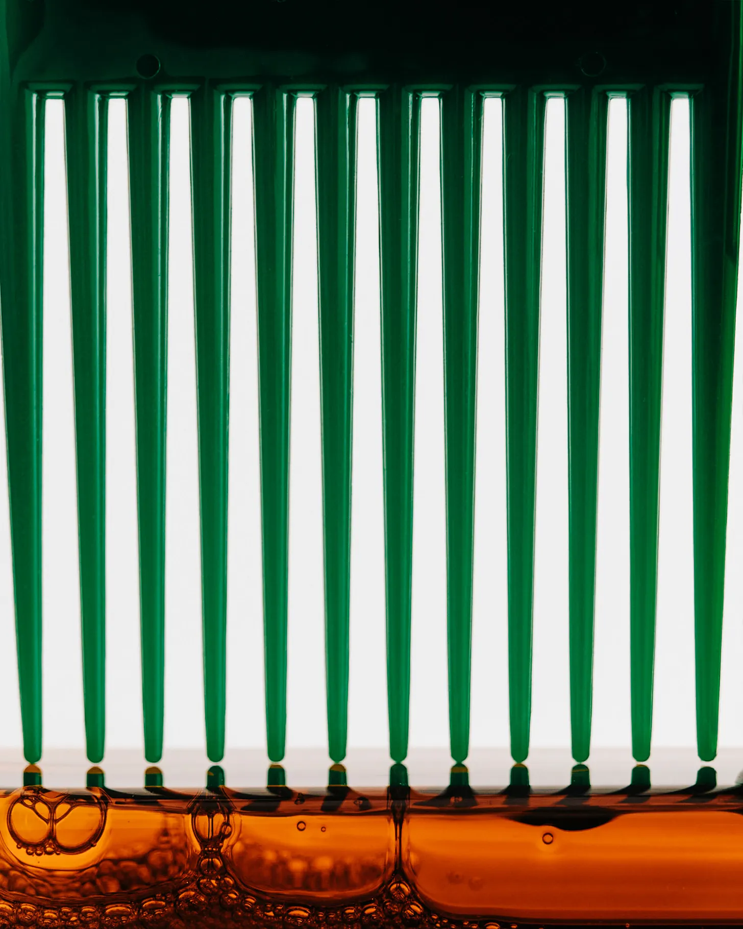 An abstract close-up of vertical green glass-like comb above a band of amber liquid filled with small bubbles, set against a bright white background. 