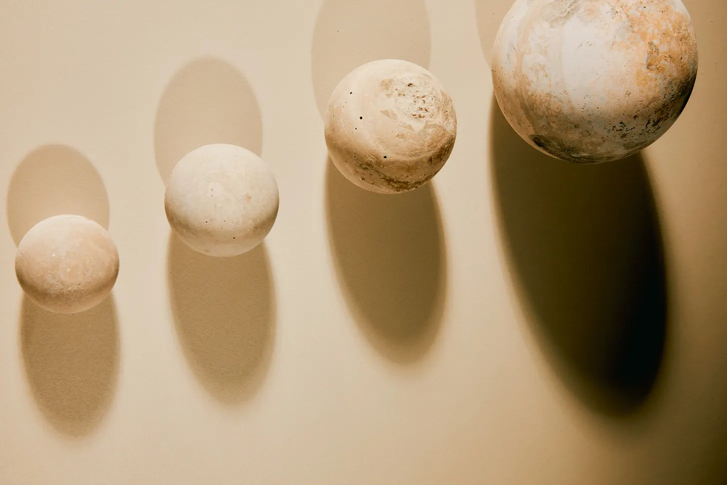 Four pale stone spheres of increasing size are arranged across a warm beige surface, each casting a long oval shadow. Their weathered, textured surfaces and strong directional lighting create a minimal sculptural composition.