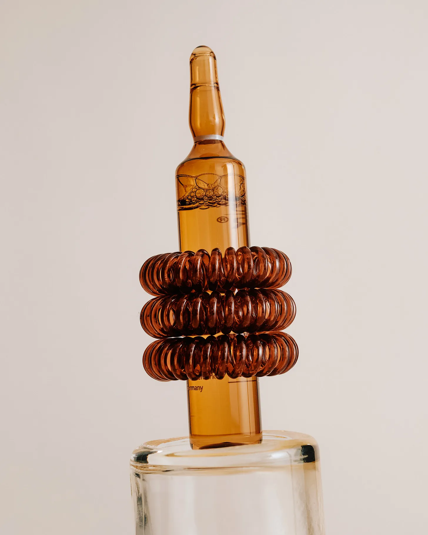A glass ampoule filled with amber liquid stands upright on a clear glass base, wrapped around the middle with three stacked translucent brown spiral rings. Tiny bubbles are visible inside the liquid against a pale neutral background. 