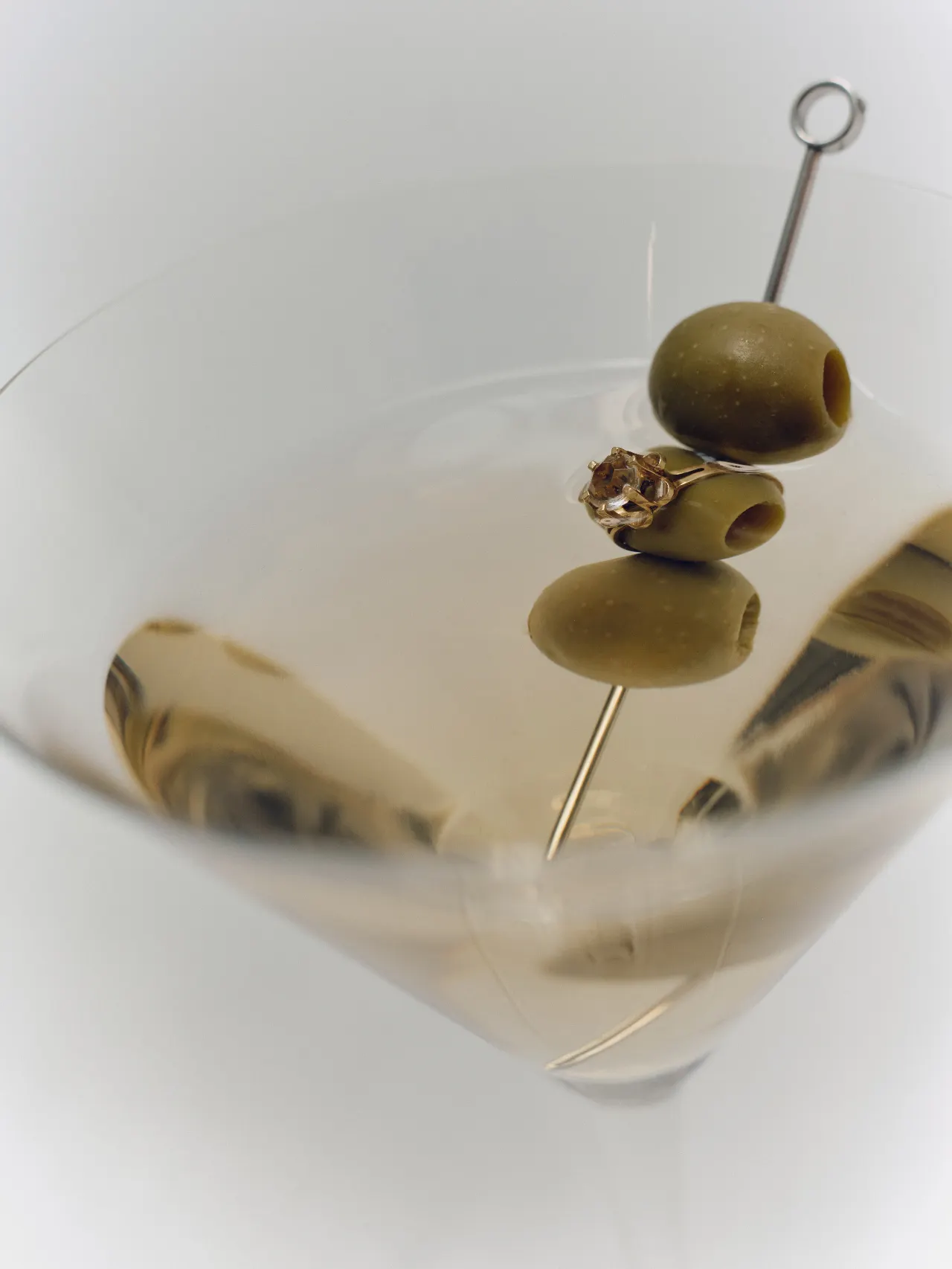A close-up of a clear martini glass filled with a pale, nearly colorless drink. Three green olives are stacked vertically on a thin silver metal cocktail pick, with the top olive near the rim and the lower two descending into the glass. An engagement ring with a prominent diamond stone is placed around the middle olive, turning the garnish into a jewelry-like focal point. The reflective surface of the drink creates mirrored images of the olives, ring, and metal pick. 
