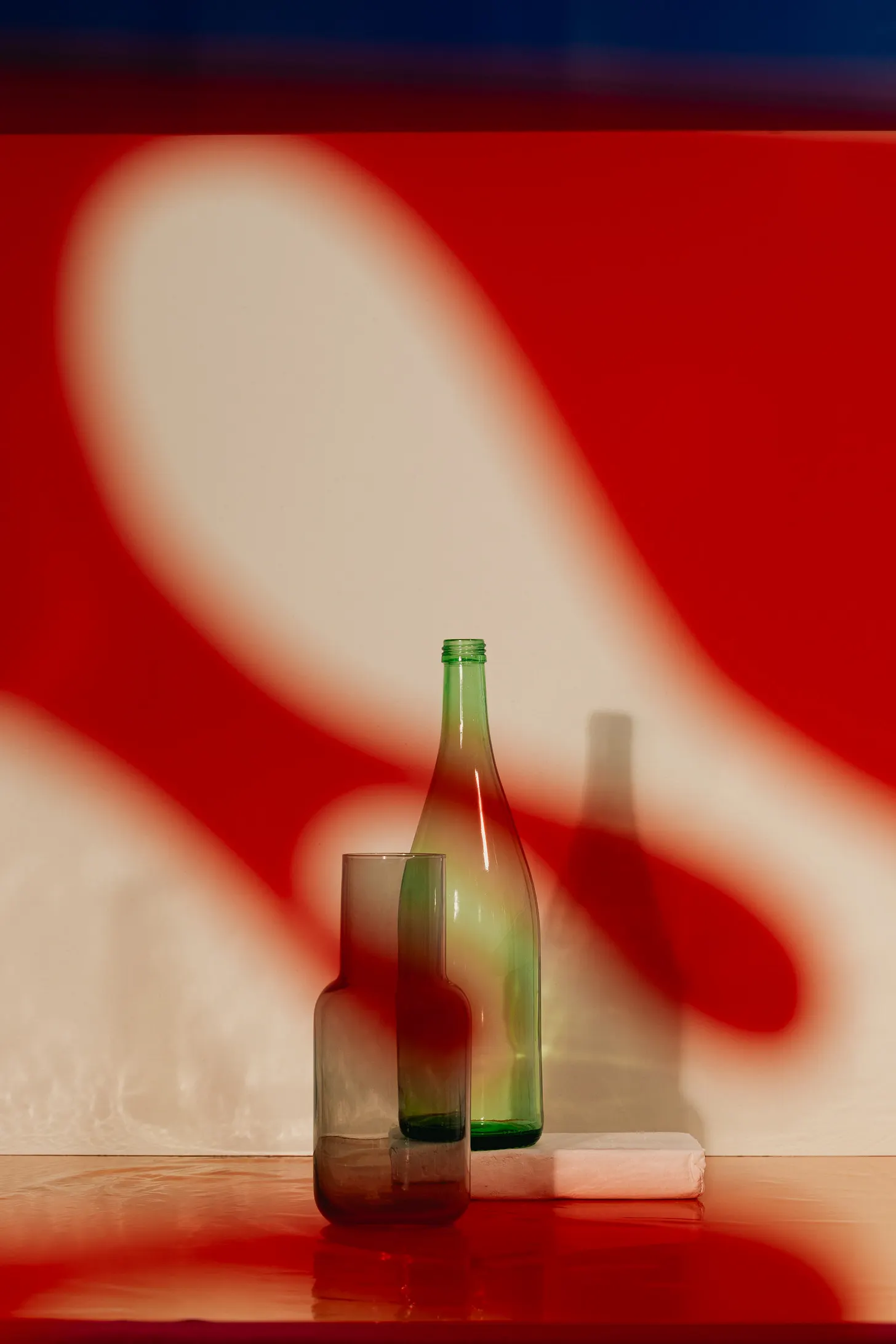 An editorial still life of two translucent glass bottles arranged on a reflective surface beside a neatly folded white cloth. A tall green bottle stands behind a shorter smoky brown bottle, both catching warm highlights from the light. Large, soft-edged red and cream light shapes sweep across the wall and tabletop, creating dramatic abstract shadows around the bottles. A narrow band of deep blue runs along the top of the image, adding contrast to the saturated red lighting and pale background.