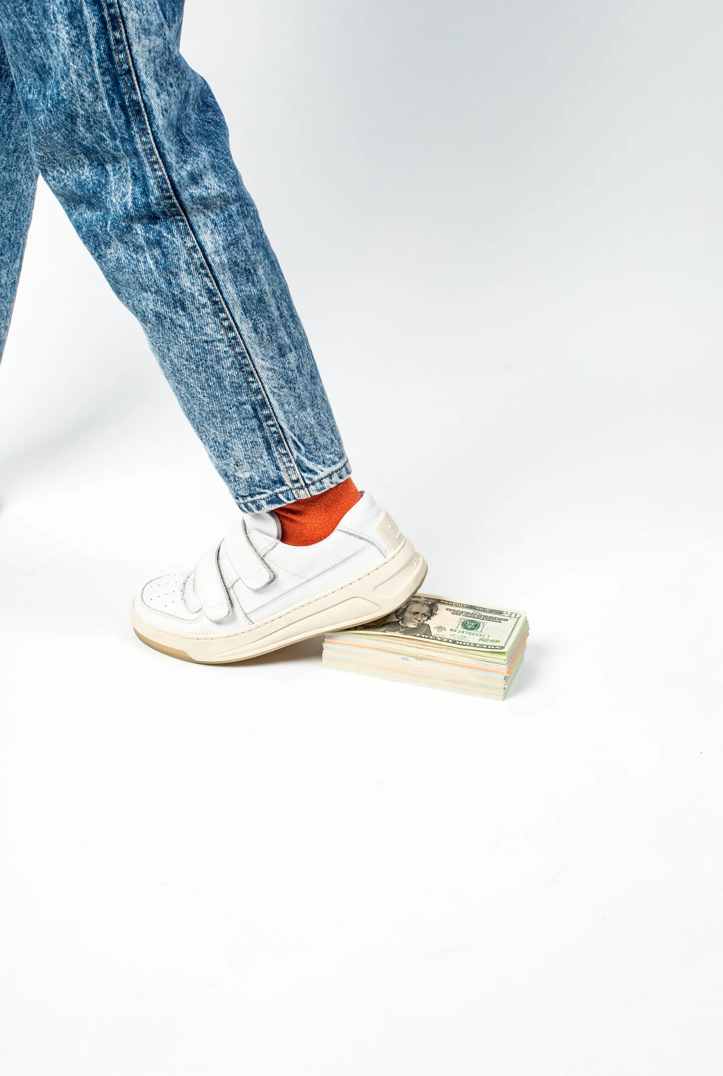 A person wearing faded blue jeans, orange socks, and white sneakers rests one foot against a thick stack of U.S. dollar bills in a walking motion on a clean white background. 