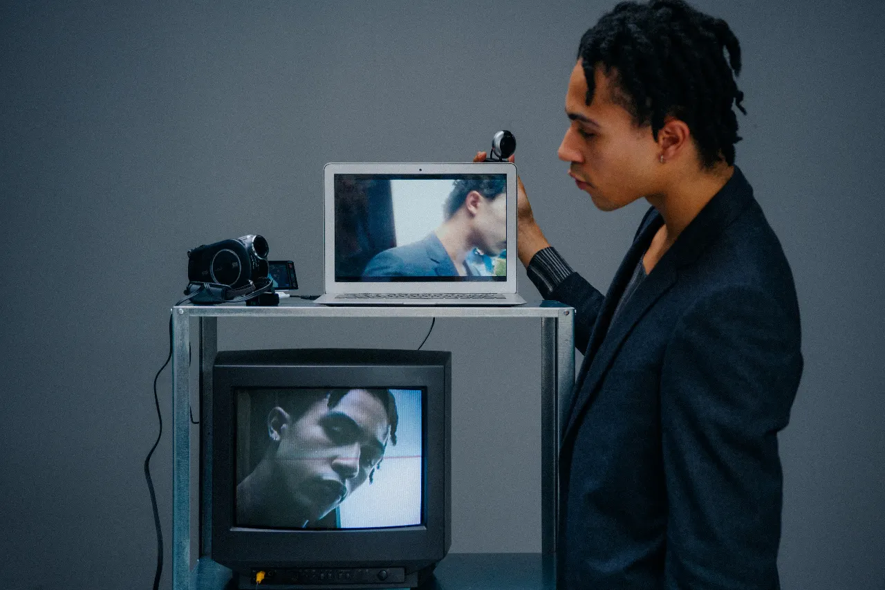 A person stands beside a laptop and television displaying different close-up views of their face, with video cameras positioned around the screens.