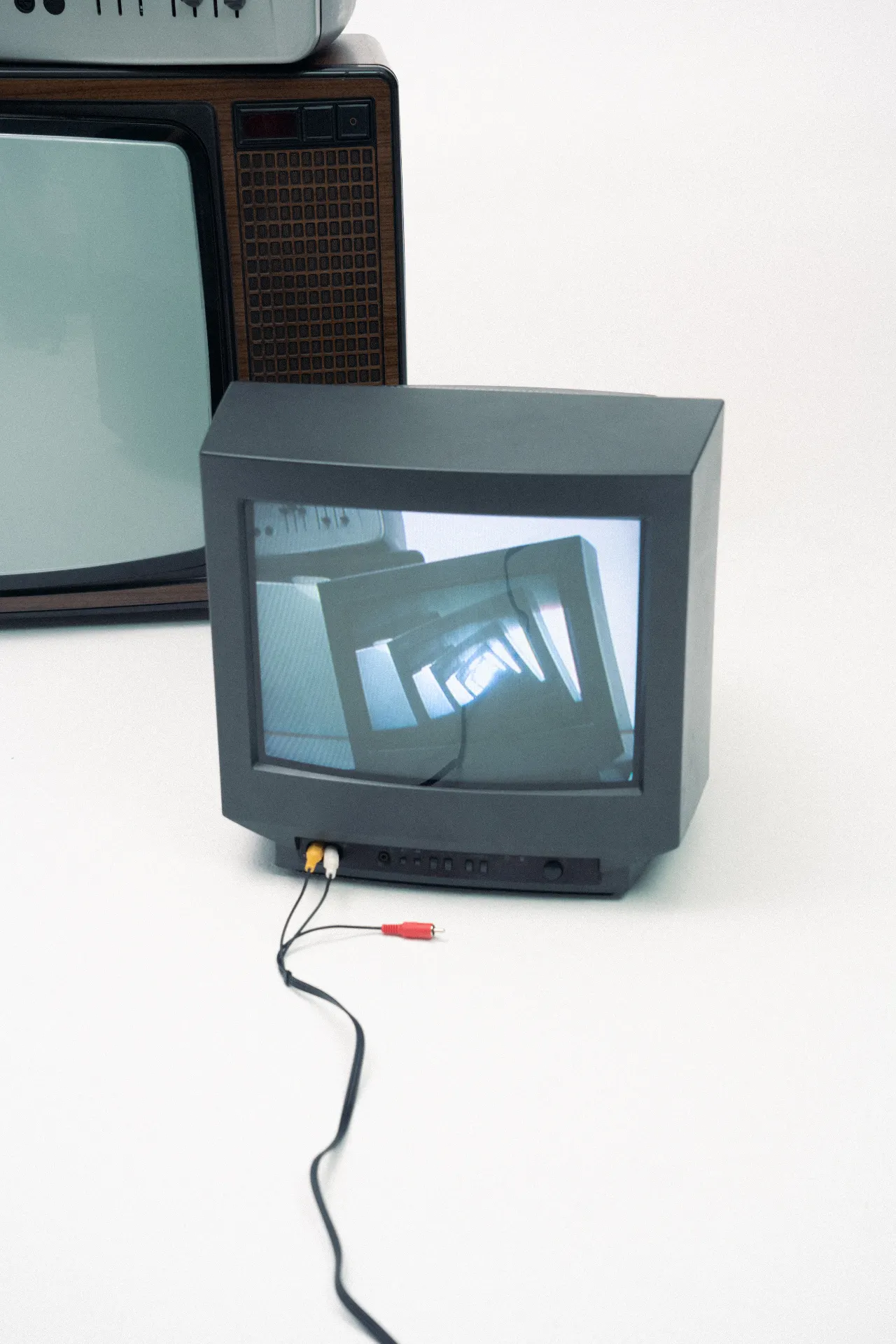 A small gray CRT television sits on a white surface with red, yellow, and white RCA cables plugged into the front, while a larger vintage television stands behind it. The smaller screen reflects a repeating tunnel-like image.
