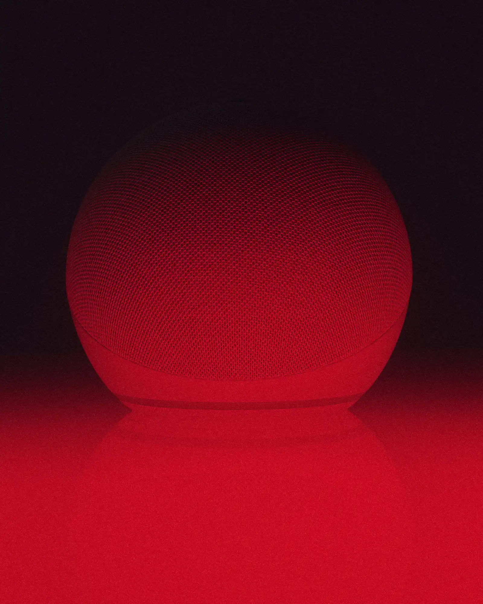 Close-up of a rounded mesh-covered listening device or speaker illuminated entirely in deep red light, with a soft shadow beneath it against a dark background.