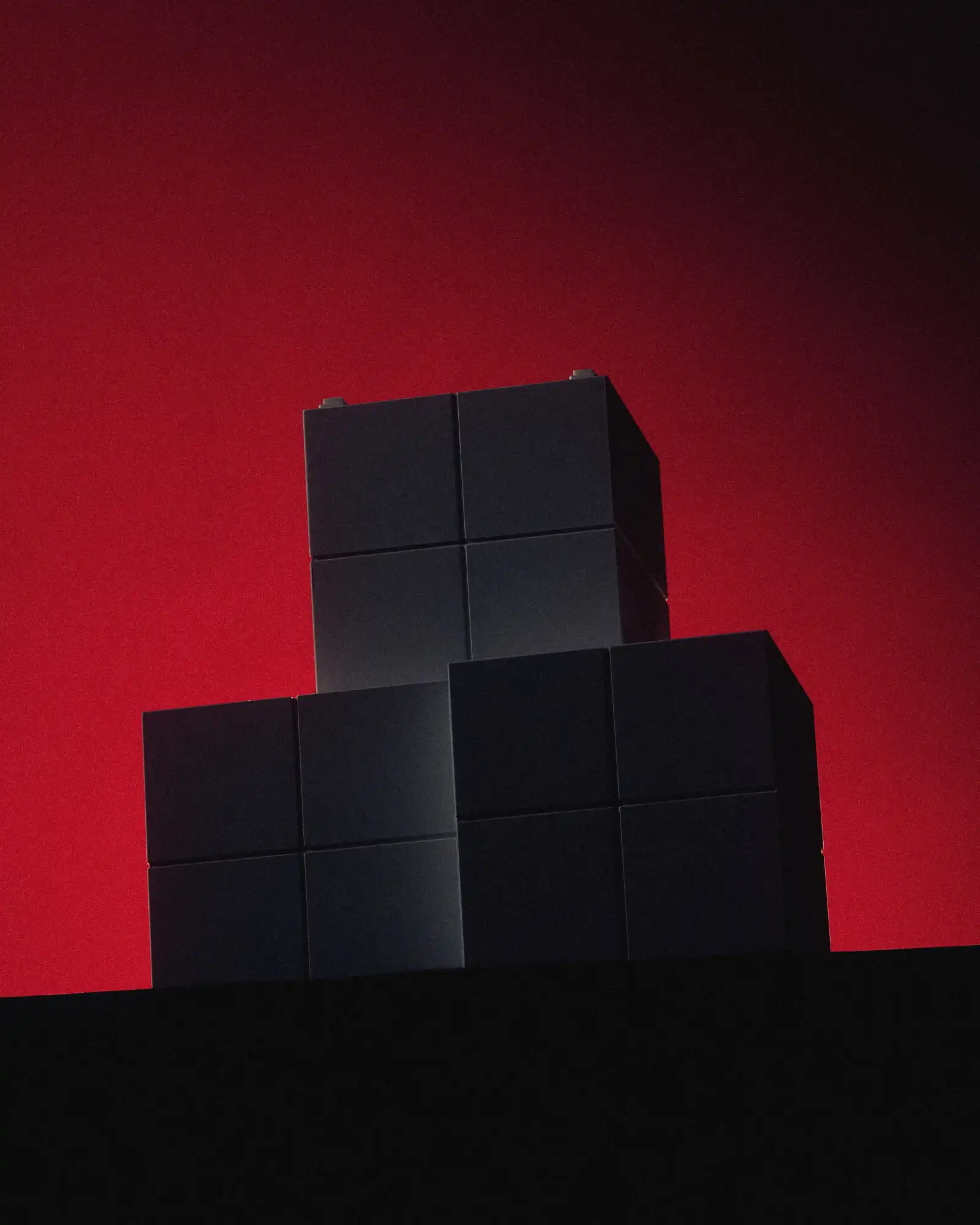 A stack of matte black cube-shaped blocks arranged like a stepped tower against a dramatic red-to-black gradient background. The geometric forms are mostly in shadow, creating a stark, sculptural silhouette.