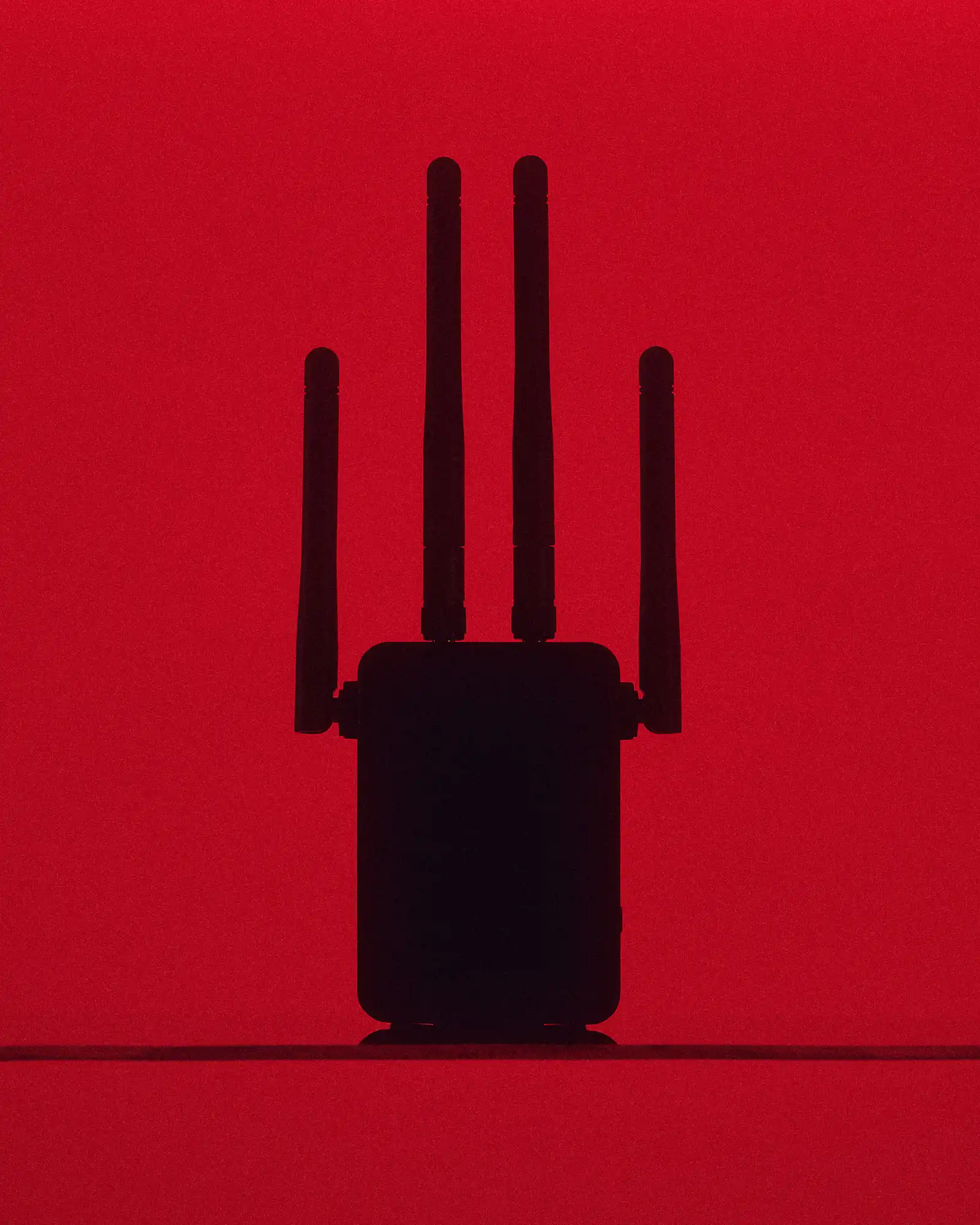 A black wireless router or communication device with four tall antennas appears in silhouette against an intense red background, creating a stark, minimal technological image.