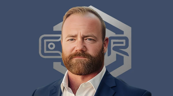 A man with a full reddish beard in a navy blazer and white open-collar shirt.