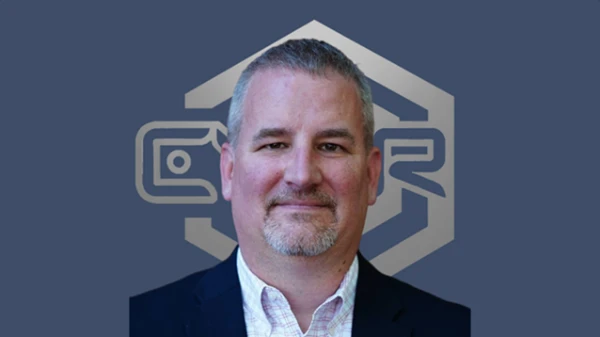 A man with grey hair and a short grey beard in a navy blazer and checked shirt, smiling slightly.