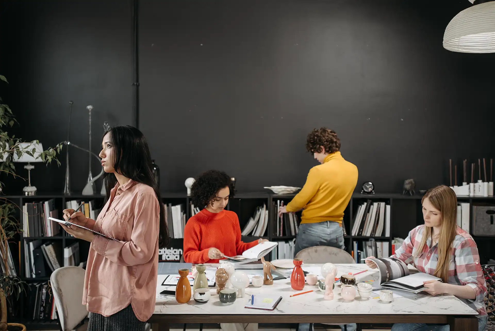 Four young adults in a modern workspace with a marble table displaying various ceramic vases and decorative objects. One woman in a pink shirt is standing and writing in a notebook, another woman in an orange sweater is reading a book, a man in a yellow sweater is selecting a book from a black bookshelf, and a woman in a plaid shirt is seated and reviewing a book. The background features a black wall and shelves filled with books and decor items.