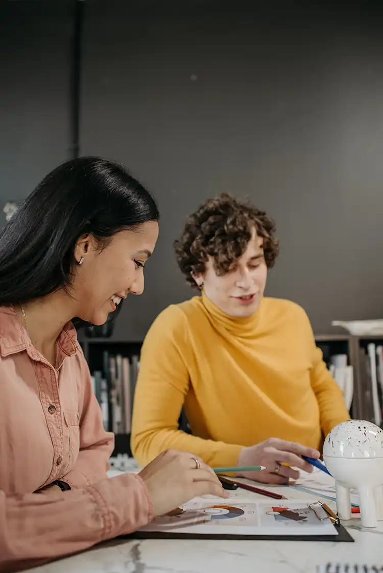 Two young adults sitting at a table reviewing a chart; one person in a pink shirt smiles while holding a pencil, and the other in a yellow turtleneck gestures with a pen.