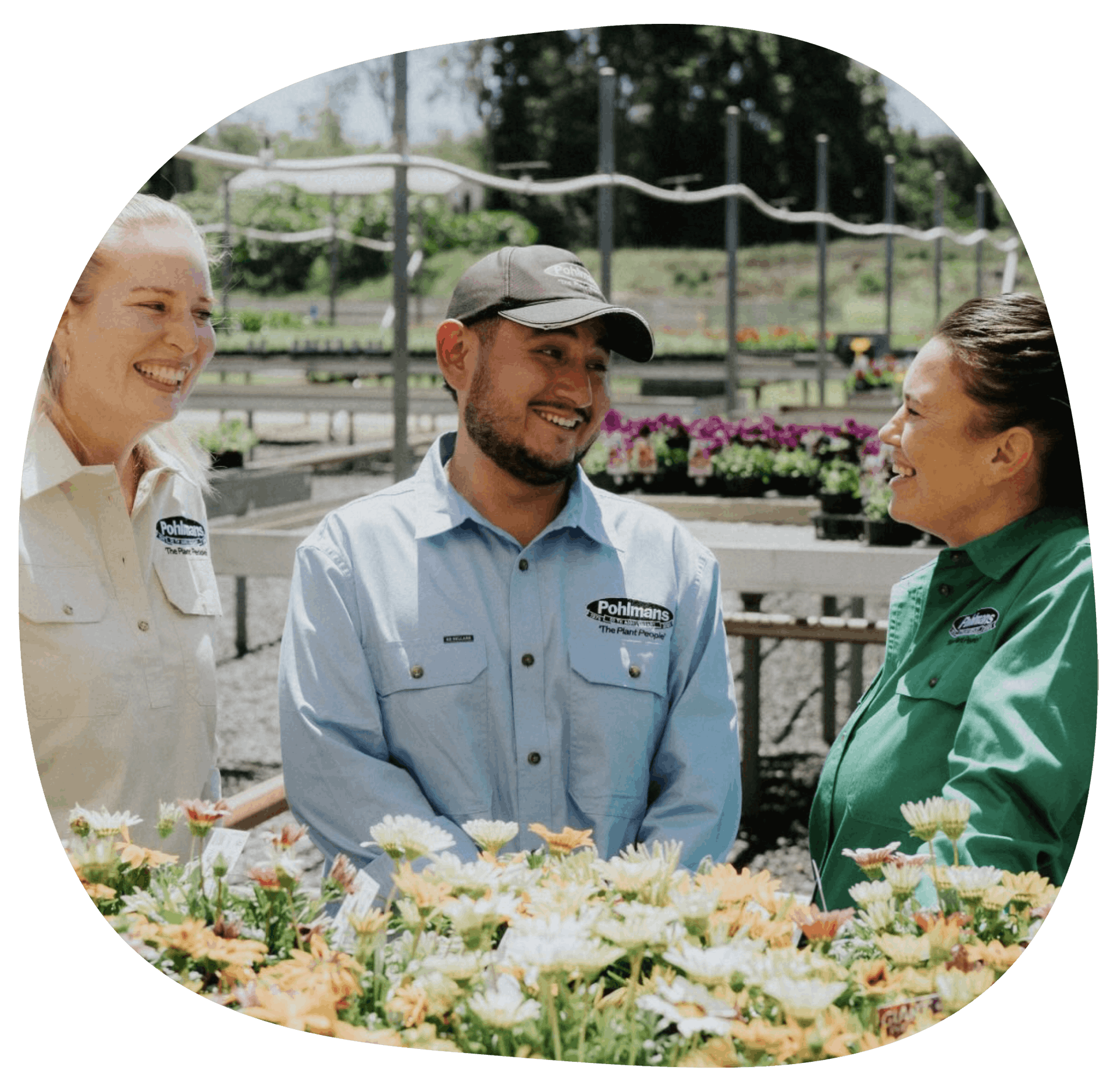 two women stand on either side of a man, they are all chatting and standing in front of a tray of flowers grown by Pohlmans.