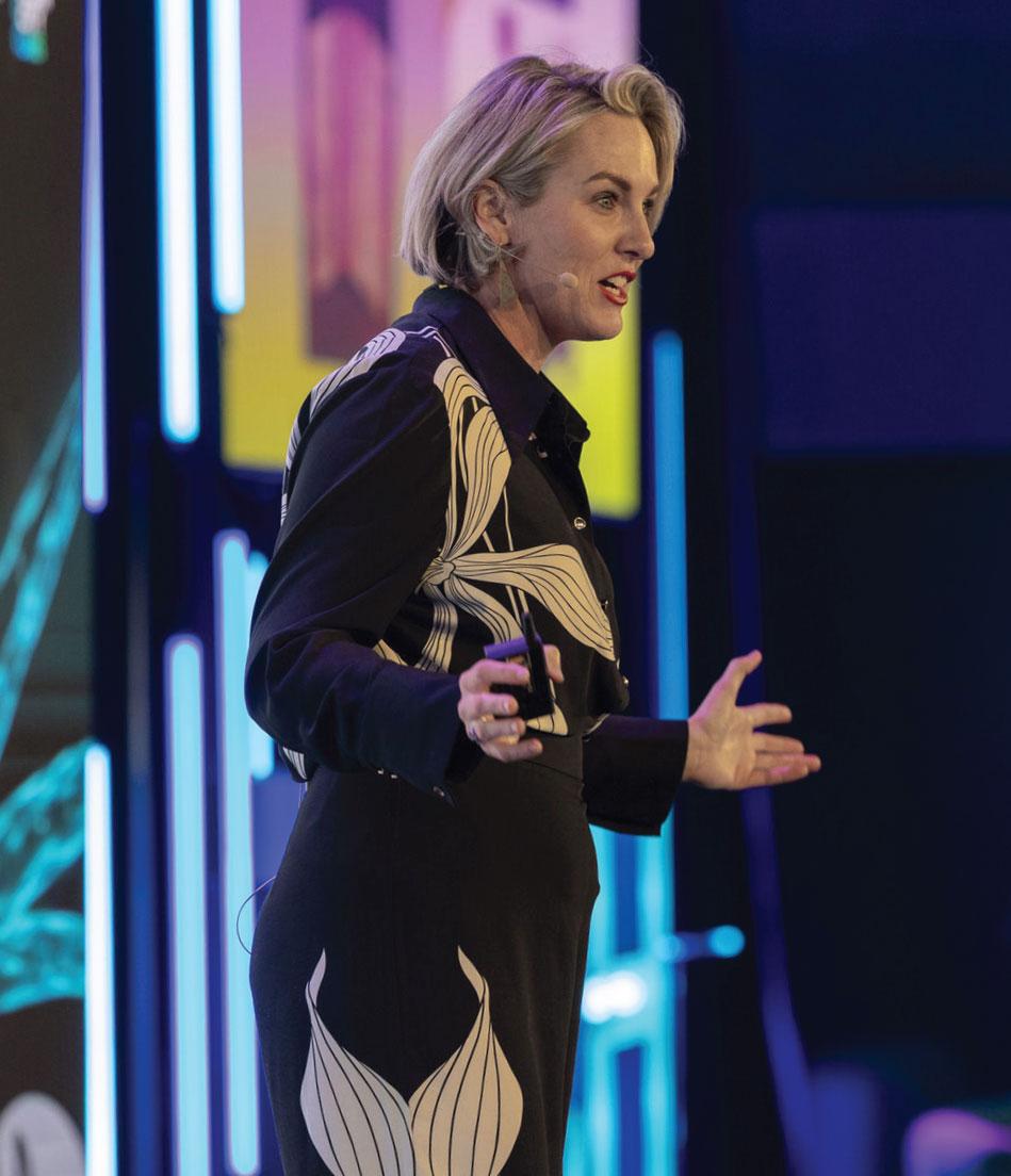 a blonde woman in a collared shirt wears a headset, while she talks to a large crowd at a conference.