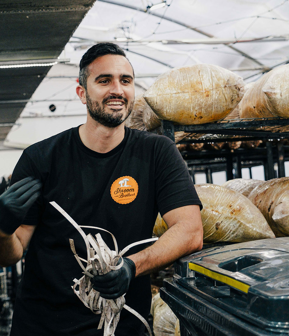 a bearded man smiling in front of shelves of growing mushrooms