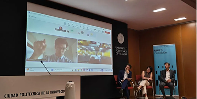 Three people seated on chairs on a stage in front of a large screen displaying a virtual meeting with multiple participants.