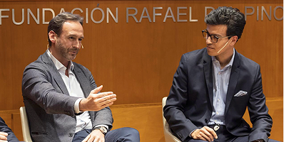 Two men seated and talking during an event, one gesturing with his hand, the other listening attentively, with a wooden background displaying partial text.