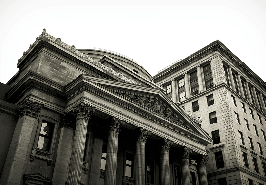 Low-angle view of a classical building with tall Corinthian columns and a triangular pediment, next to a rectangular building with multiple windows and columns at the roofline.