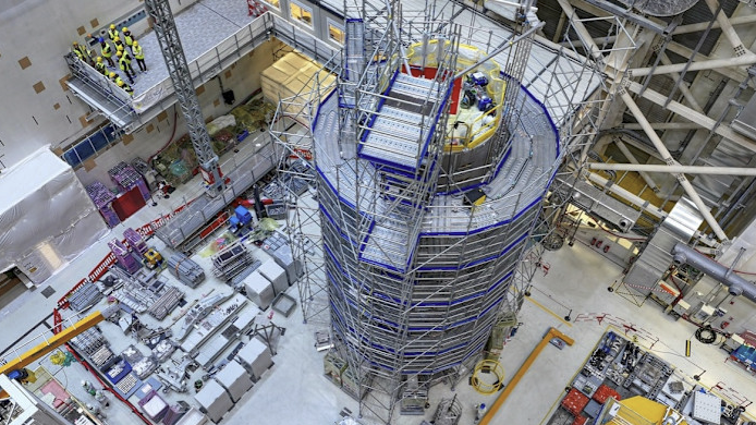 Overhead view of a large industrial construction site with a tall cylindrical structure surrounded by scaffolding and various equipment and materials organized on the floor.