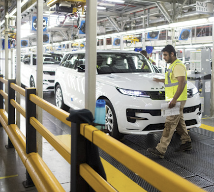 Factory worker in a yellow safety vest inspecting a white Range Rover vehicle on an assembly line inside a modern automotive factory.