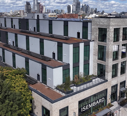 Modern multi-story building with brick and gray panel facade, rooftop terraces with greenery, and large windows, set against a city skyline with skyscrapers under a partly cloudy sky.