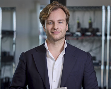 Man with light brown hair wearing a dark blazer over a white shirt, standing indoors with blurred shelves and equipment in the background.