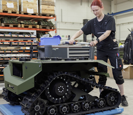 Person with red hair wearing black work clothing adjusting equipment on a green, tracked robotic vehicle inside a workshop with shelves of wooden boxes and tools in the background.