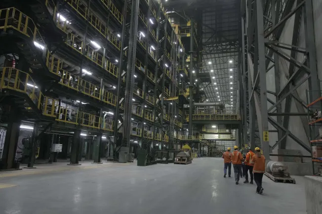 Interior of a large industrial facility with tall metal structures and walkways on the left, and six workers wearing orange jackets and yellow helmets walking on the spacious floor to the right.