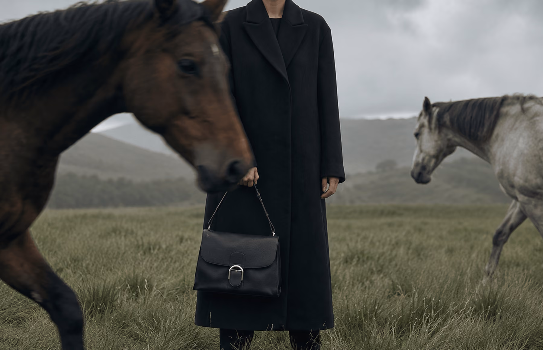 Woman in a long black coat holding a leather handbag beside a horse