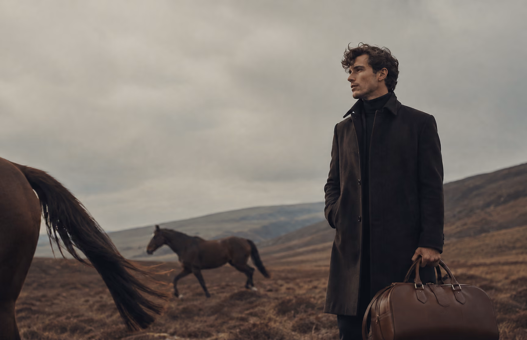 Man in a long dark coat holding a leather bag in a windswept field, a horse behind him