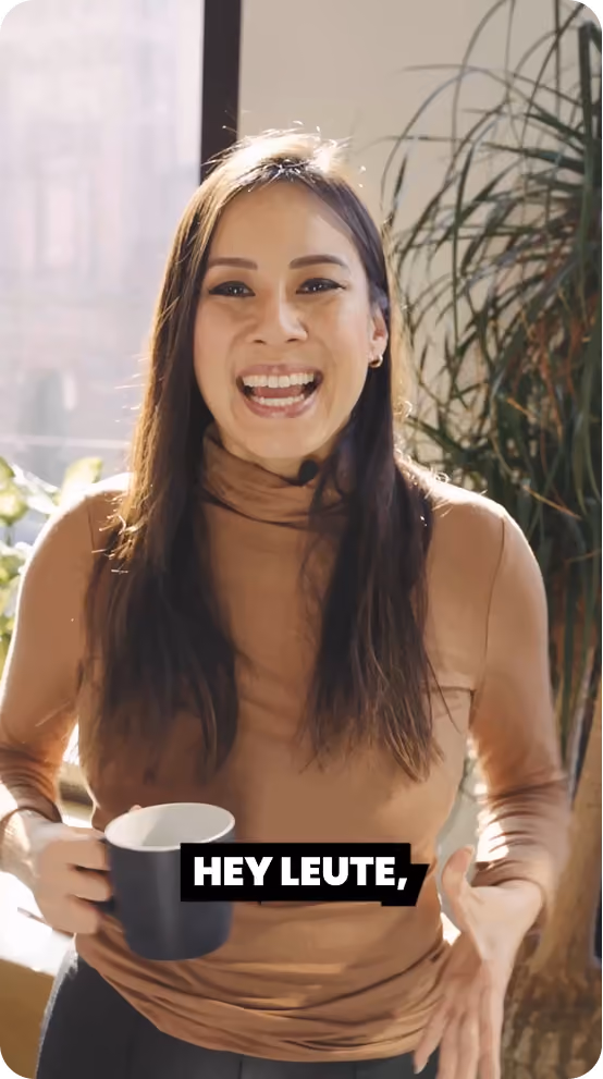 Lächende Frau mit langen braunen Haaren und braunem Rollkragenpullover hält eine schwarze Tasse, im Hintergrund Pflanzen und Fensterlicht.