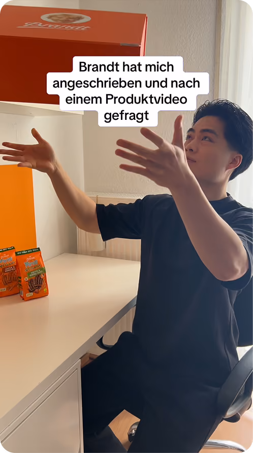 Ein junger Mann in schwarzem T-Shirt sitzt an einem Schreibtisch und hebt beide Hände in die Luft, während eine rote Brandt-Verpackung über ihm schwebt und zwei weitere Packungen auf dem Tisch stehen. Text in der Bildmitte lautet: „Brandt hat mich angeschrieben und nach einem Produktvideo gefragt“. Das Zimmer ist hell mit einem Fenster im Hintergrund.